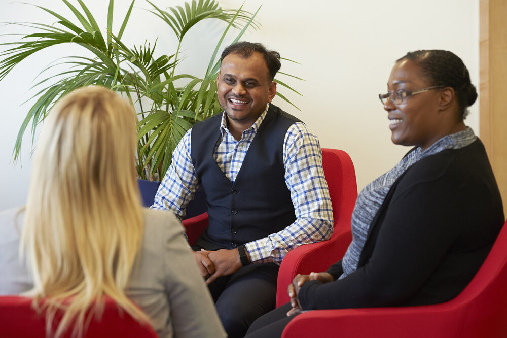 Three office employees sitting facing each other in red chairs, smiling and laughing at each other.